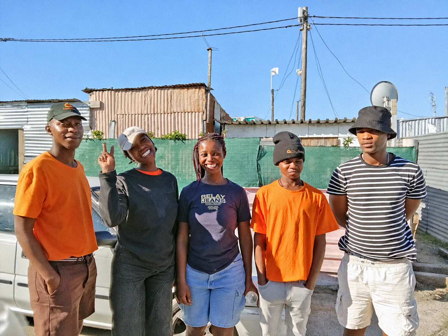 Five young people, two in DigiDeeds caps and orange T-shirts, standing together in front of homes in their neighbourhood