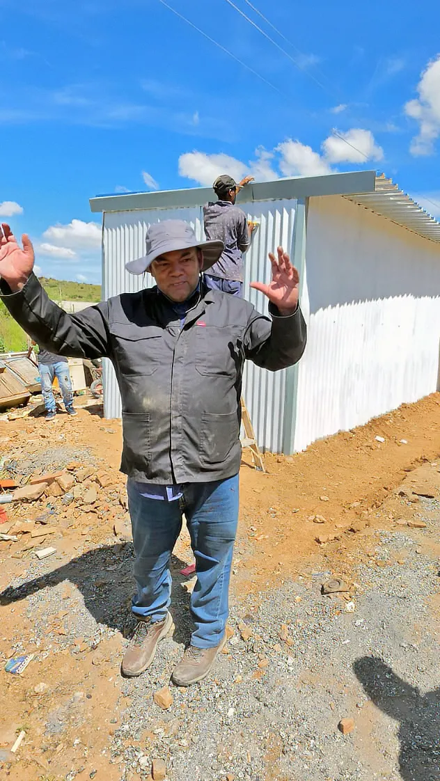 A builder in a sunhat with his arms spread in front of a newly clad home, as a colleague works on the roof