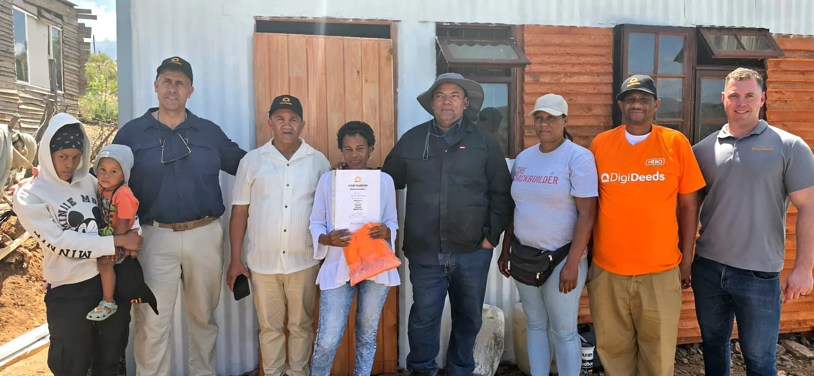 A resident holding her DigiDeed, with her family and the DigiDeeds and Shackbuilder teams, in front of her new home