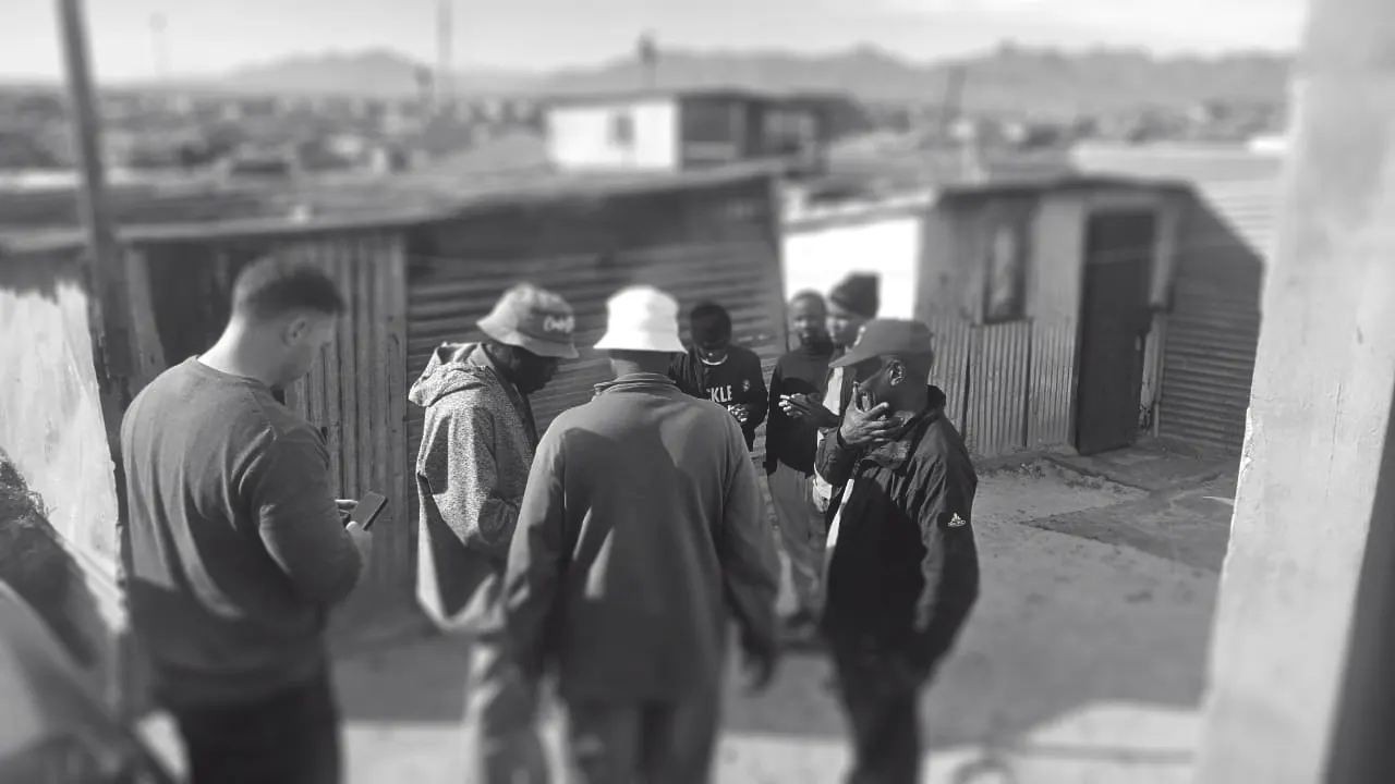 Black-and-white photograph, softly blurred, of a group of people standing together between homes in their neighbourhood