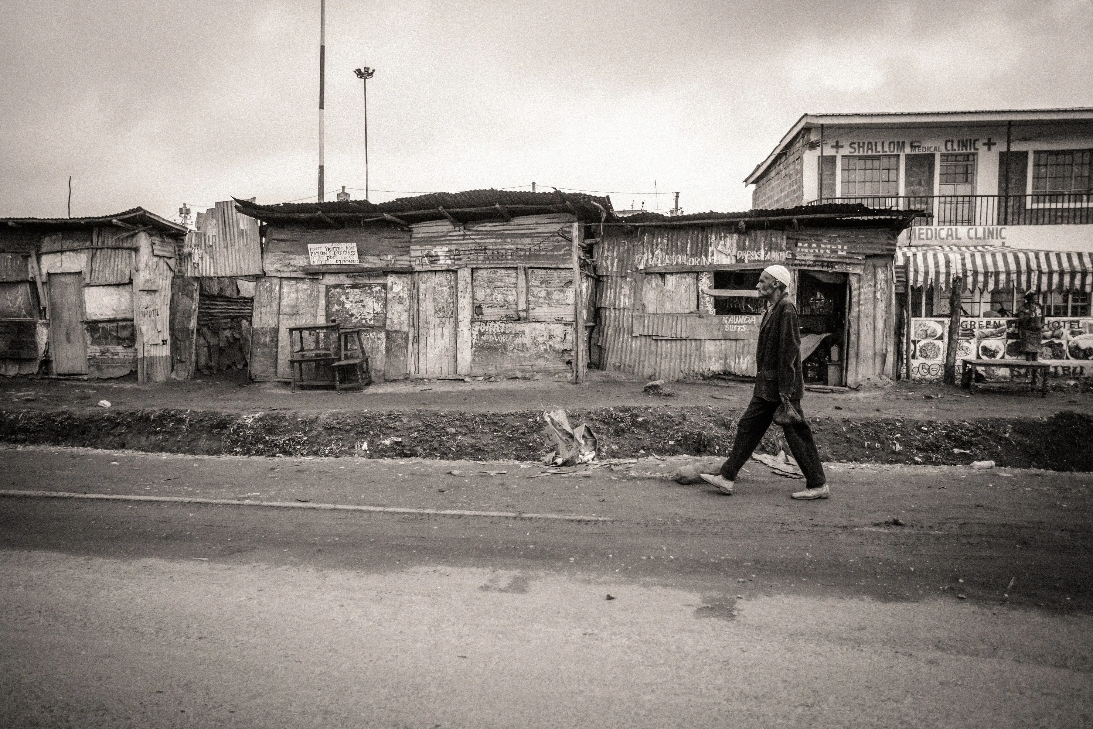 Sepia-toned photograph of a man walking along a road past a row of corrugated-iron shops and homes